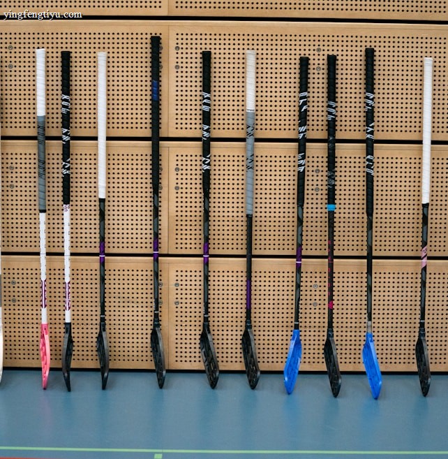 A row of floorball sticks, leaning vertically against a perforated wooden wall. The sticks have white handles and various colored blades, including pink, blue, and black shades. The floor is a smooth, greyish-blue surface.