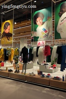 A retail store showcases clothing and sportswear on mannequins and hangers, with large vibrant posters featuring children in sports attire above. The interior displays bright garments and accessories on shelves and racks.