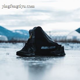 A pair of black sports shoes and a black bag with the brand name 'Bauer' are placed on an icy surface. The background features blurred mountains and a cloudy sky, creating a serene and somewhat cold atmosphere.