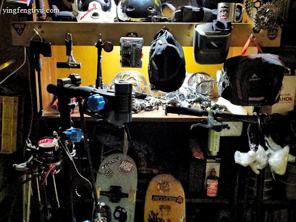 A workshop area filled with various biking and sports equipment. Several bike helmets are neatly arranged on an upper shelf. Below, there are bicycle parts, tools, and other gear. Two snowboards with graphics are leaning against the wall, and biking gear such as gloves and hats are scattered around. The area is lit with a bright overhead light, highlighting the equipment.