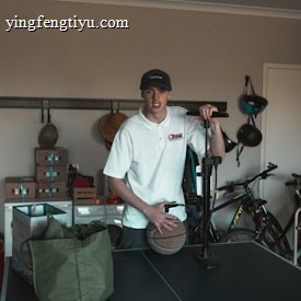 A person wearing a white shirt and a cap is standing in a room with various items. The person is holding a basketball and seems to be next to an air pump. In the background, there are bicycles and shelves with boxes. A green bag is placed on a black table, which appears to be a table tennis table.