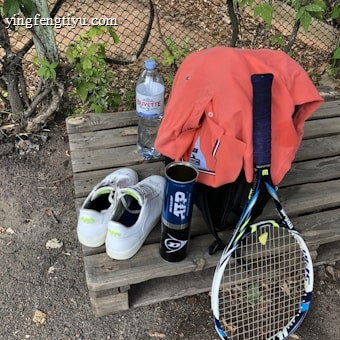 A tennis racket lies on a wooden bench alongside a pair of white tennis shoes with green accents. A blue canister, possibly for tennis balls, is next to an orange shirt draped over a black bag. A large bottle of water sits nearby, with a wire fence and greenery in the background.